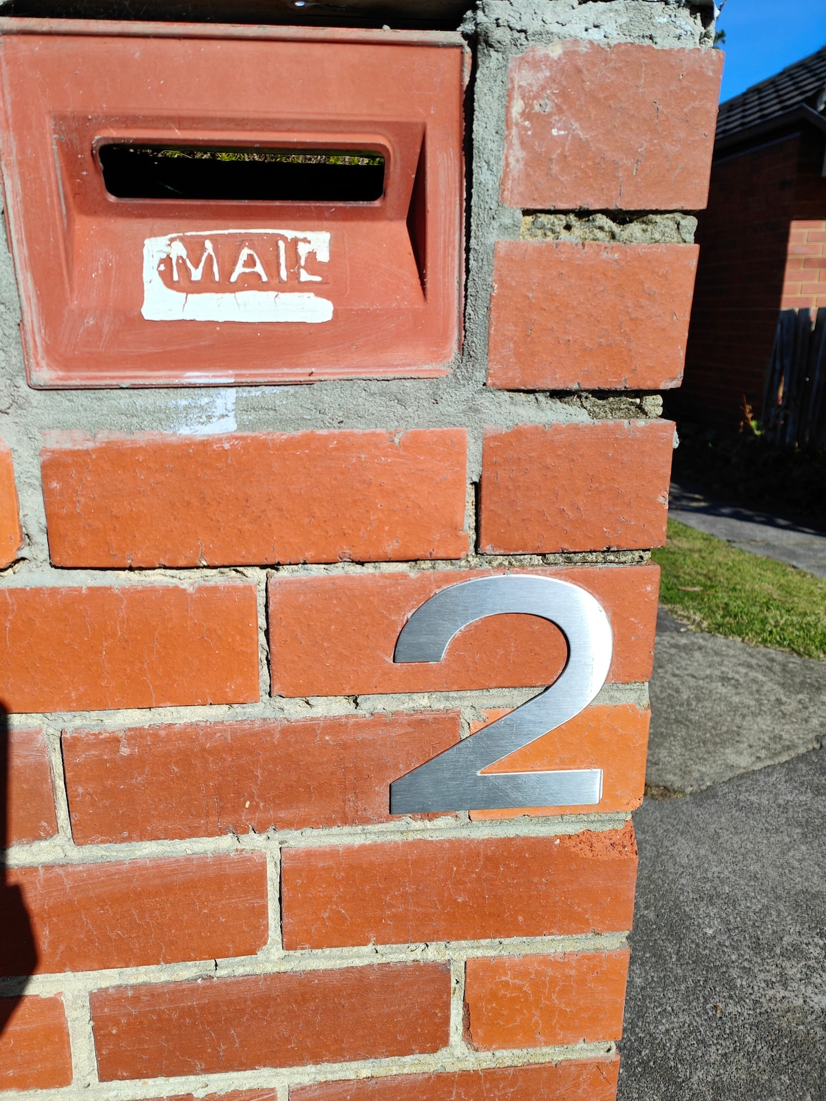 A close-up view of a brick wall features a mailbox in a reddish hue, labeled 'MAIL' in white paint. The bold silver numeral '2' is prominently displayed beside the mailbox, indicating the address. Green grass is visible in the background.