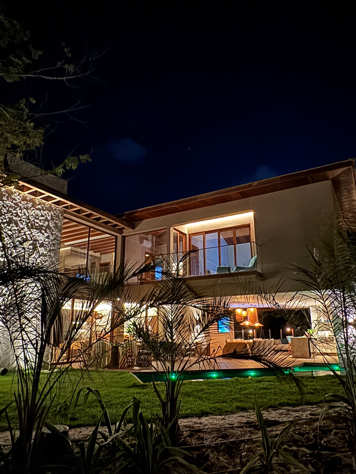 A two-story residence is illuminated at night, showcasing large glass windows that reveal a cozy interior. Lush greenery surrounds the property, and a subtle light highlights the exterior stone wall, creating a serene atmosphere amid nature.