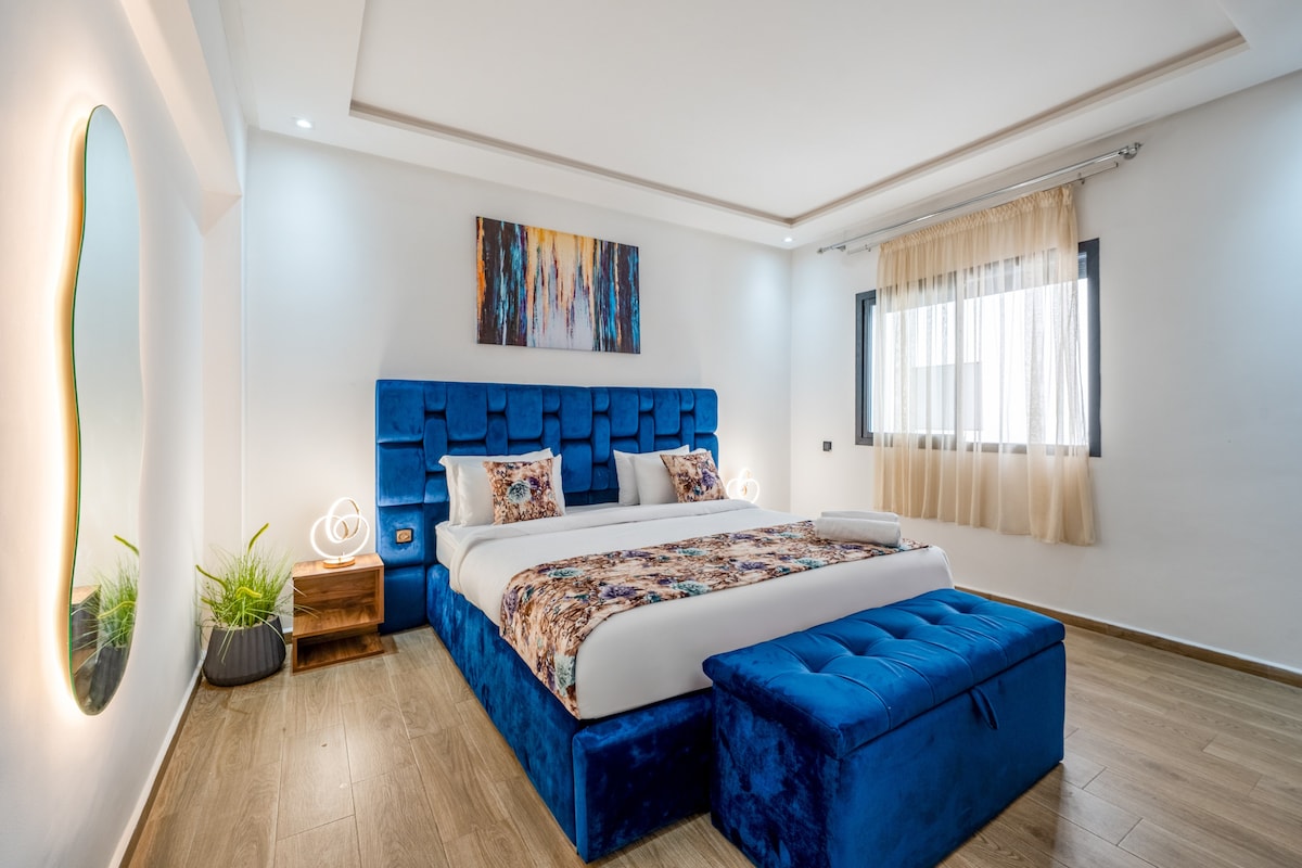 A spacious bedroom is presented with a large, soft blue bedframe and a comfortable mattress adorned with decorative pillows. Natural light filters through sheer curtains at the window, while a textured wall mirror reflects the calming environment. A stylish storage bench complements the decor.