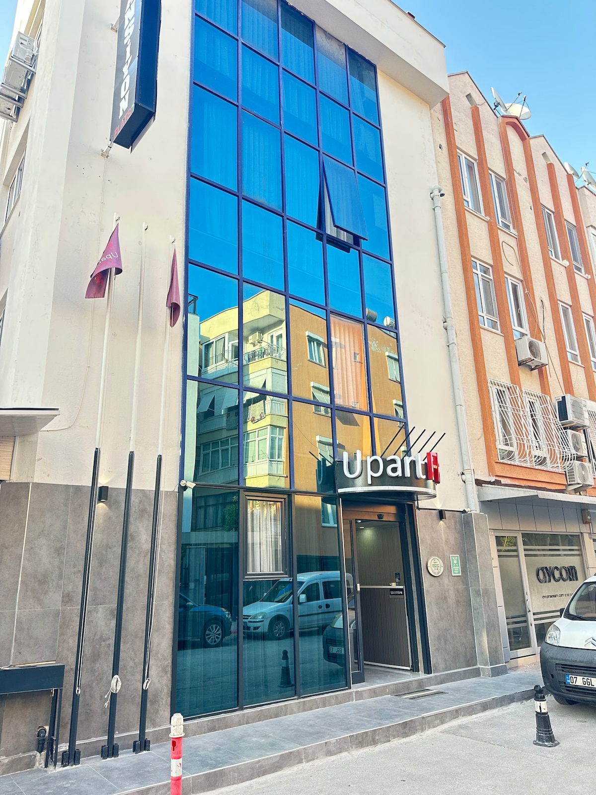 The building's modern façade is characterized by large mirror-like glass panes that reflect its surroundings. The entrance is marked by a sleek sign displaying the name 'Upart,' complemented by decorative flagpoles and minimalist architectural details.
