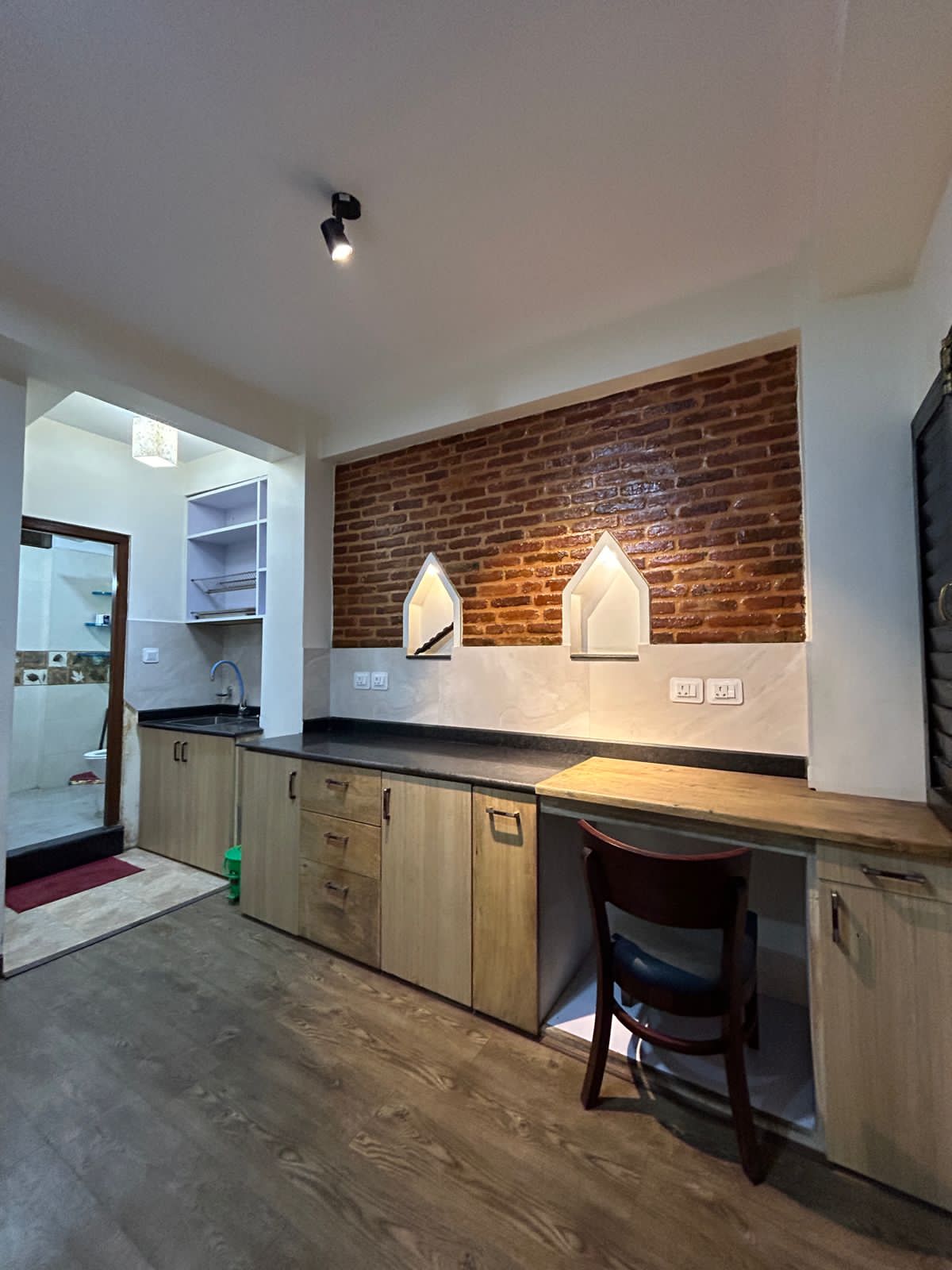 A kitchenette with wooden cabinetry features a black countertop. The wall is adorned with red brick, and two decorative alcoves provide subtle lighting. A small wooden desk with a chair is positioned against the wall, offering a functional workspace. The flooring is a light wood tone.