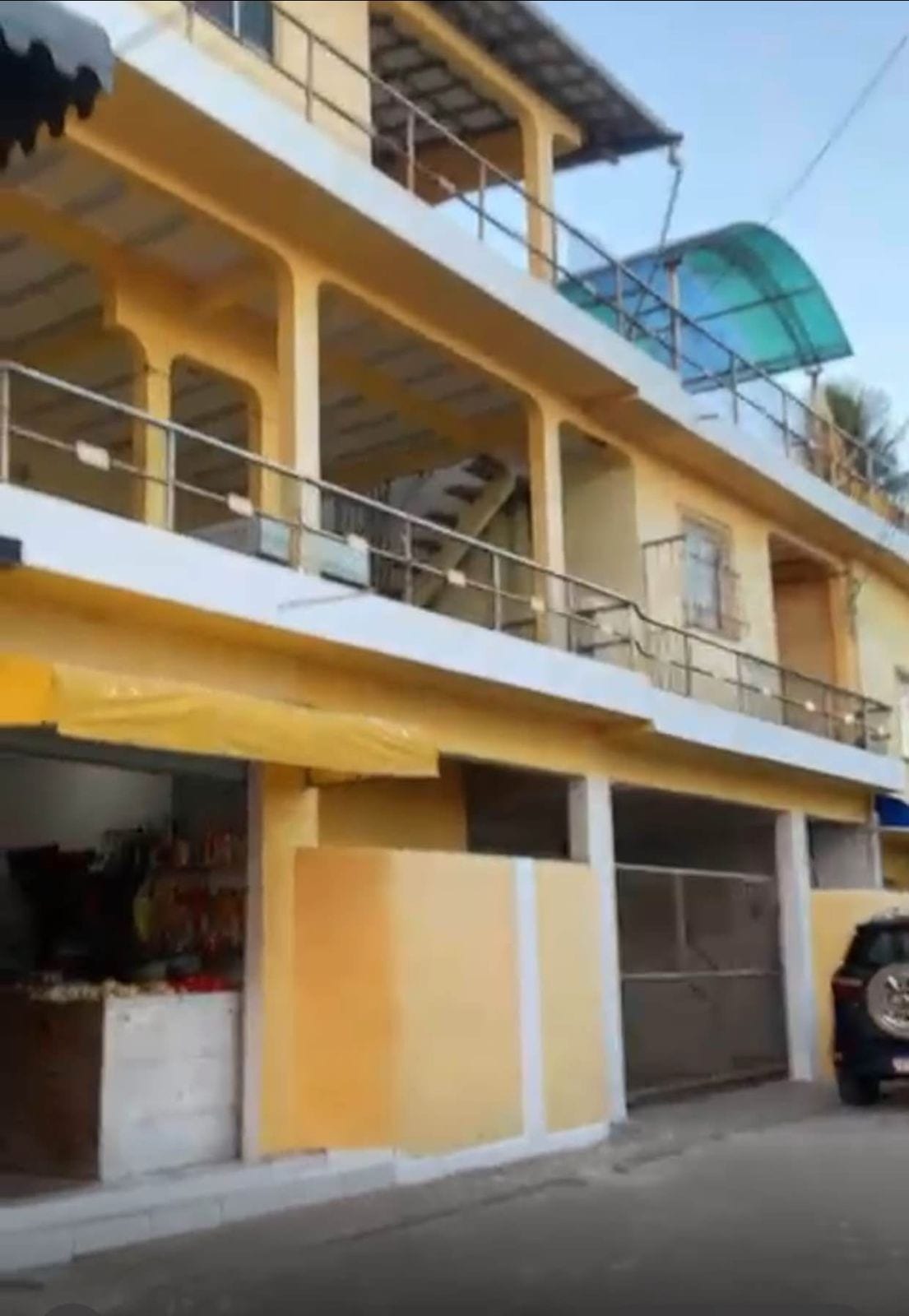 The exterior of a two-story yellow building is visible, featuring a balcony with metal railings on the upper level. A flight of stairs leads to the second floor, while a shop entrance is located on the ground floor. Nearby, a vehicle is parked beside the structure.