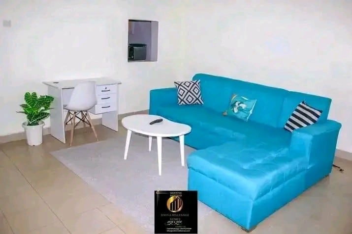 A modern living area features a light blue sectional sofa with accent pillows, positioned beside a small white coffee table on a neutral rug. A compact desk with a chair is situated against the wall, accompanied by a decorative plant in a white pot.