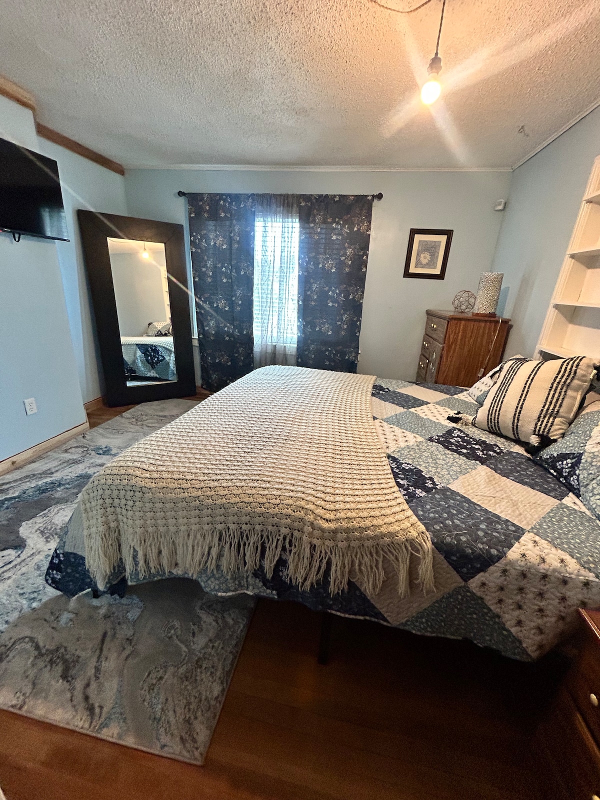 A cozy bedroom features a spacious bed adorned with a patterned quilt and a soft throw. A large mirror reflects the room’s decor, while soft light filters through a window, partially covered by a patterned curtain. Warm wooden elements enhance the inviting atmosphere.