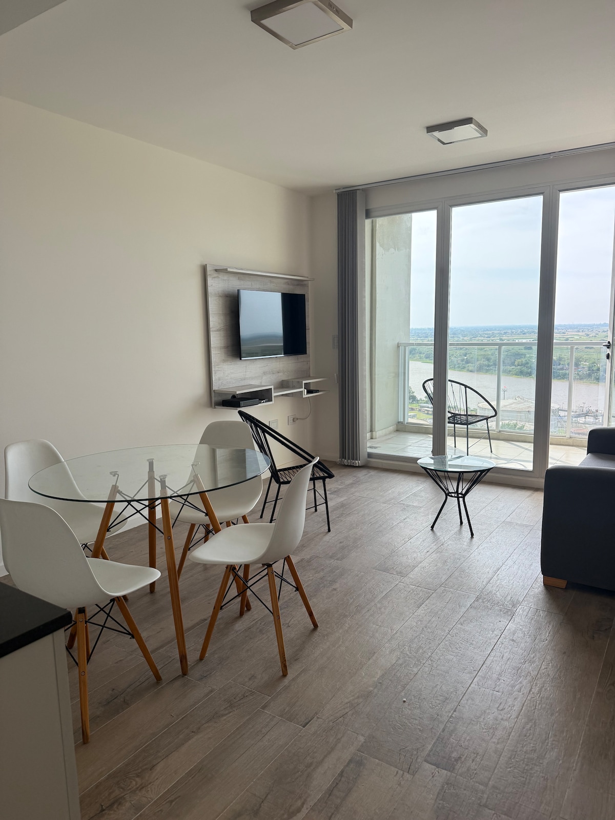 A dining area is set with a round glass table surrounded by four white chairs. A wall-mounted TV is visible above a console, and large windows provide a view of the outdoors, bringing ample natural light into the room.