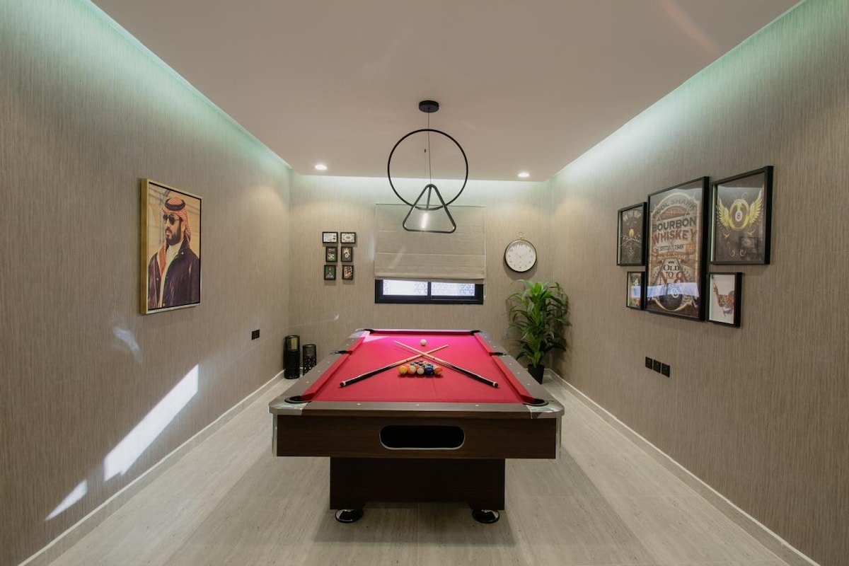 A game room is showcased, featuring a vibrant red billiard table positioned in the center. Modern light fixtures provide ambient illumination, while framed artwork and photos are displayed on the walls. A small green plant adds a touch of nature to the space.