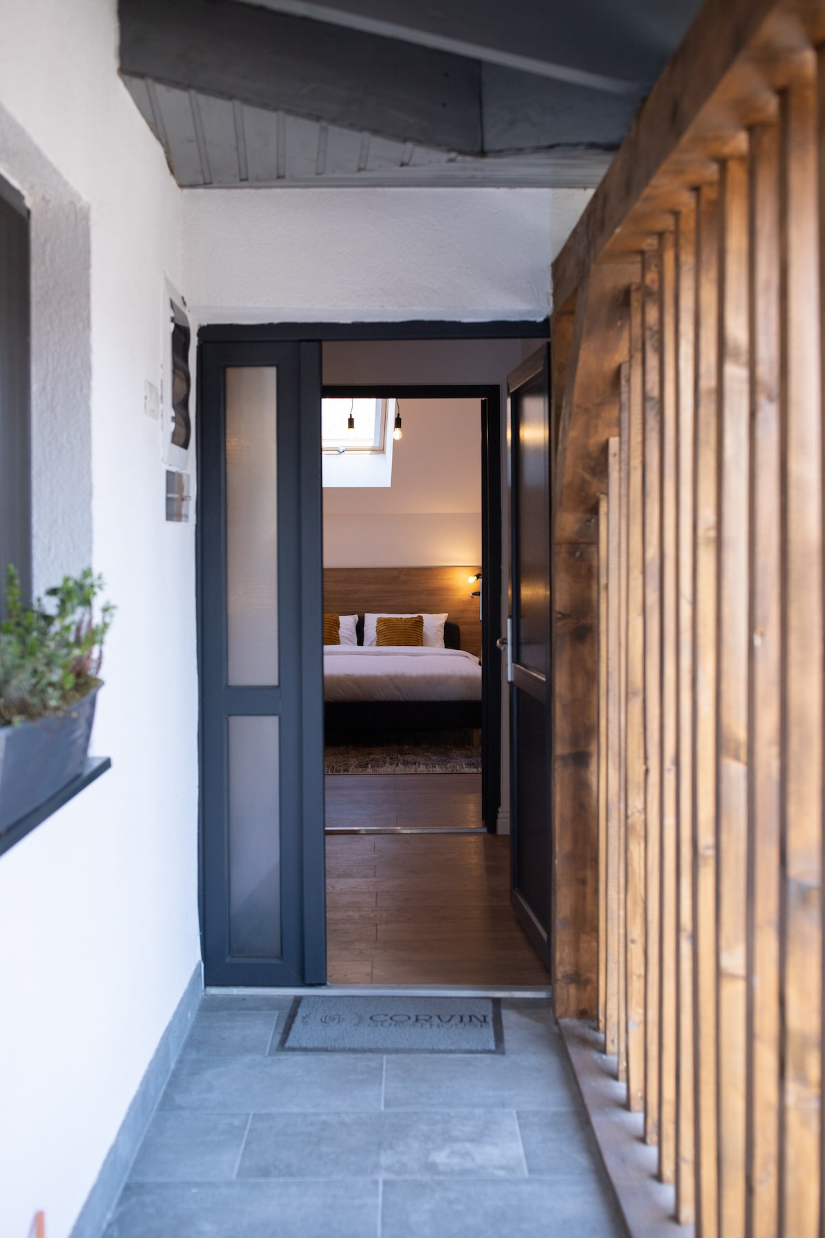 The entryway is framed by a wooden railing and features a sleek dark door that opens to reveal a glimpse of a cozy bedroom. Natural light filters into the space, highlighting muted colors and inviting décor.