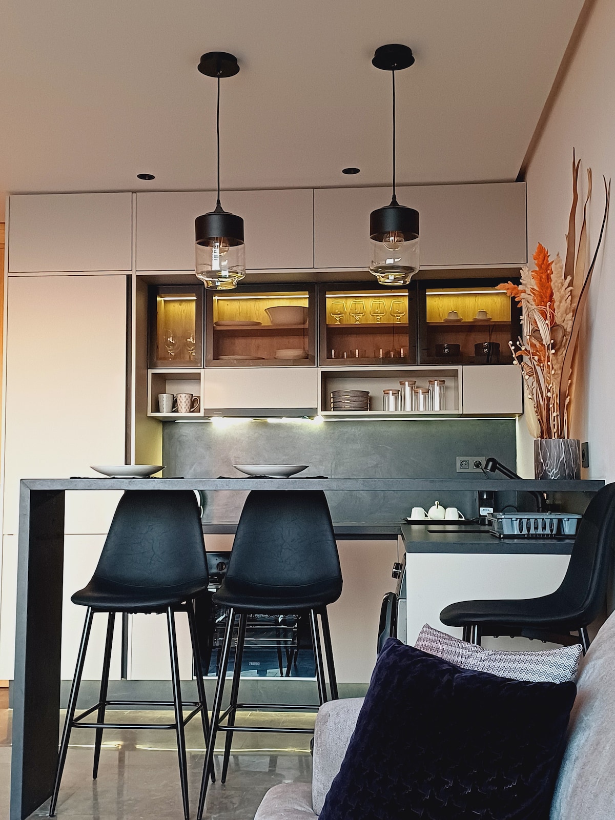 A modern kitchen area is visible, with sleek cabinetry and ambient lighting highlighting glass dishware on open shelves. Two high stools are positioned at a black bar counter, creating a space for dining or socializing. Elegant decorative elements, including dried plants, add a touch of warmth.
