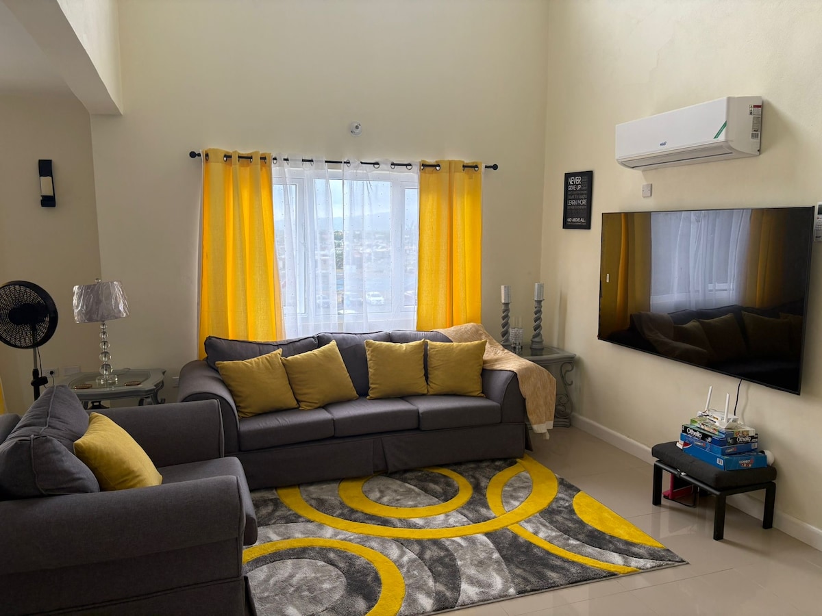 A cozy living area is presented with a gray sectional sofa adorned with yellow accent pillows. A circular area rug in gray and yellow adds warmth to the space. Natural light filters through sheer curtains, while an air conditioning unit and wall-mounted television enhance the modern design.