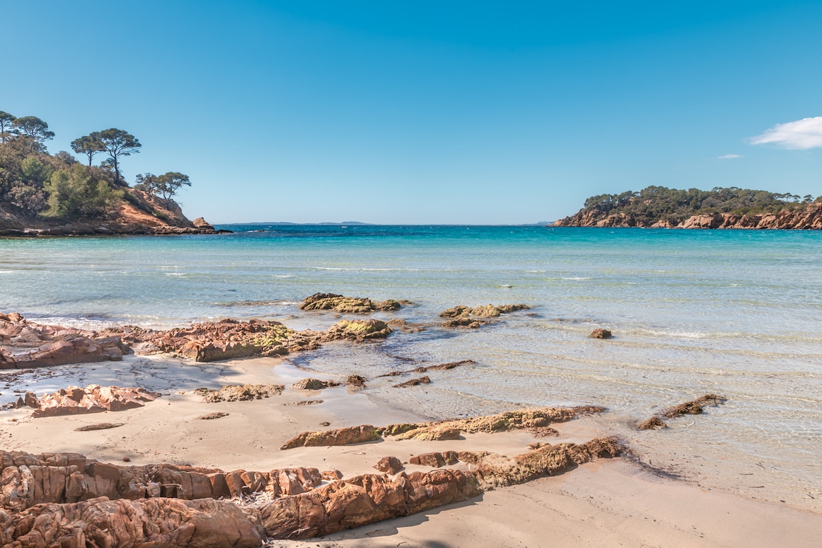 The image captures a serene beach scene with soft sand and smooth rocky formations leading into clear blue waters. Gentle waves lap at the shore, while distant islands and a clear sky complete the tranquil coastal landscape.