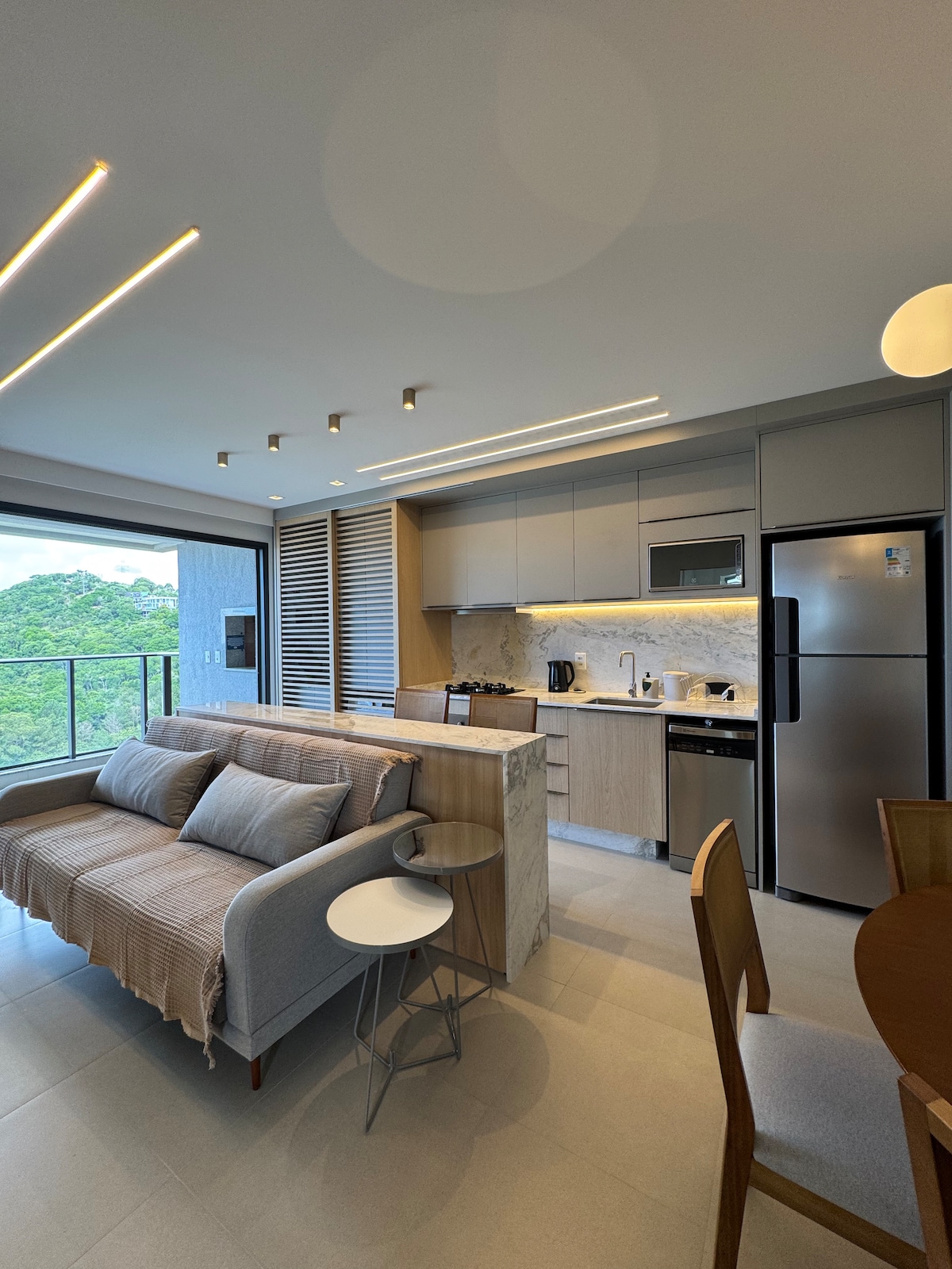 An open-concept living area is displayed, featuring a cozy sofa paired with a small, round side table. The kitchen is equipped with modern appliances, including a refrigerator and coffee maker. Natural light enters through large glass doors, leading to a balcony area with greenery views.