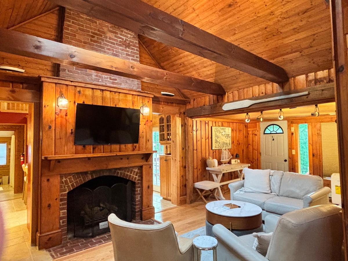 A warm living area is framed by exposed wooden beams and knotty pine walls. A brick fireplace adds rustic charm, complemented by a flat-screen television mounted above it. Light-colored couches offer a spacious seating arrangement, with a round coffee table positioned in the center. Natural light enters through nearby windows.