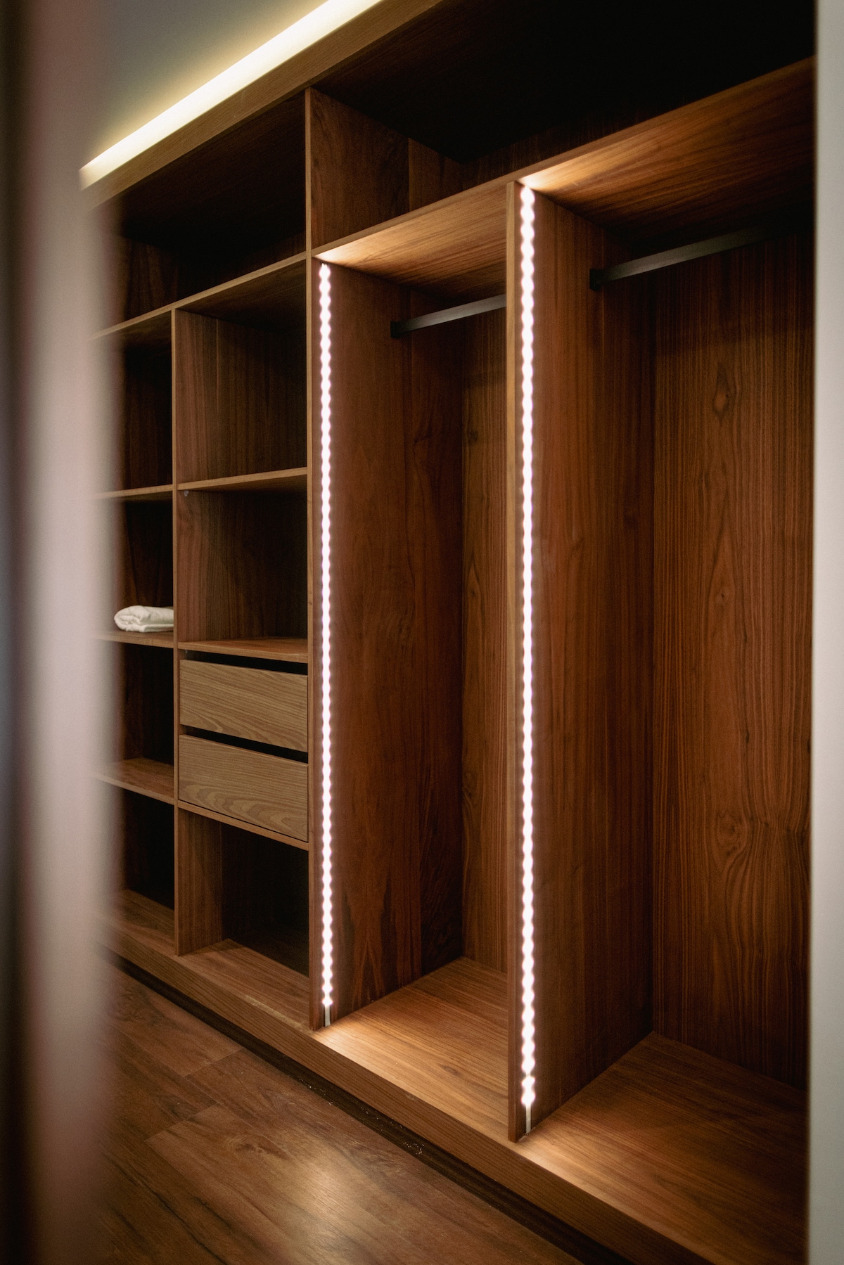 A well-organized walk-in closet is featured, constructed with sleek wooden shelving and illuminated by subtle LED lighting. The space includes open compartments for hanging clothes and drawers for storage, providing a functional and stylish area for personal belongings.
