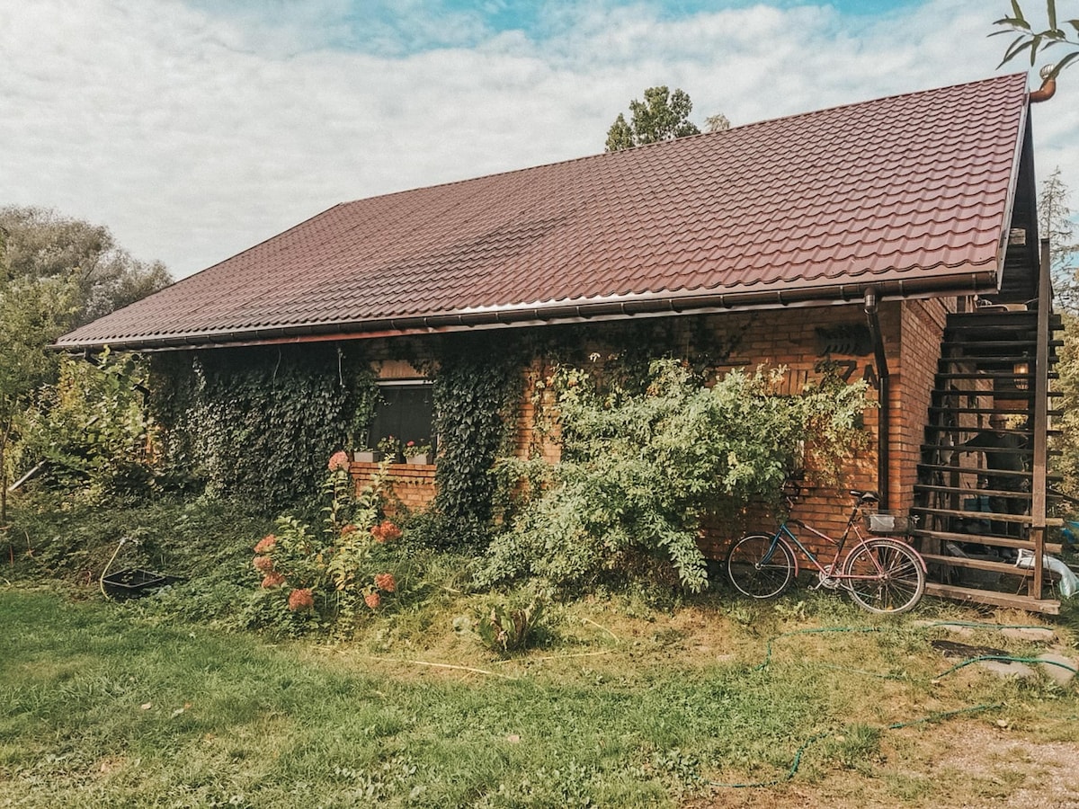 A rustic brick building is surrounded by lush greenery, with climbing plants covering one side. A bicycle is positioned near the entrance, highlighting a peaceful outdoor setting. The sloped roof and natural surroundings create a serene atmosphere, ideal for relaxation.