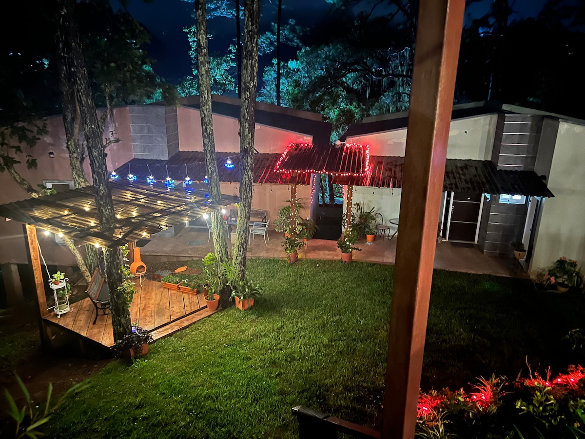 An outdoor space is illuminated by warm string lights, highlighting a wooden deck with potted plants. Two unique structures are visible in the background, showcasing varying roof styles and surrounded by greenery, contributing to an inviting atmosphere.