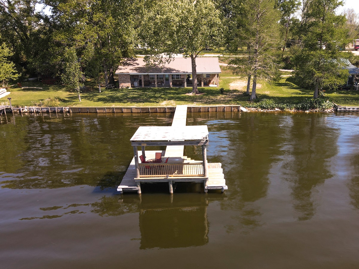 A private pier extends over calm water, featuring a small seating area under a covered roof. The renovated lakefront home is visible in the background, surrounded by greenery. Natural light reflects off the water's surface, enhancing the serene setting.
