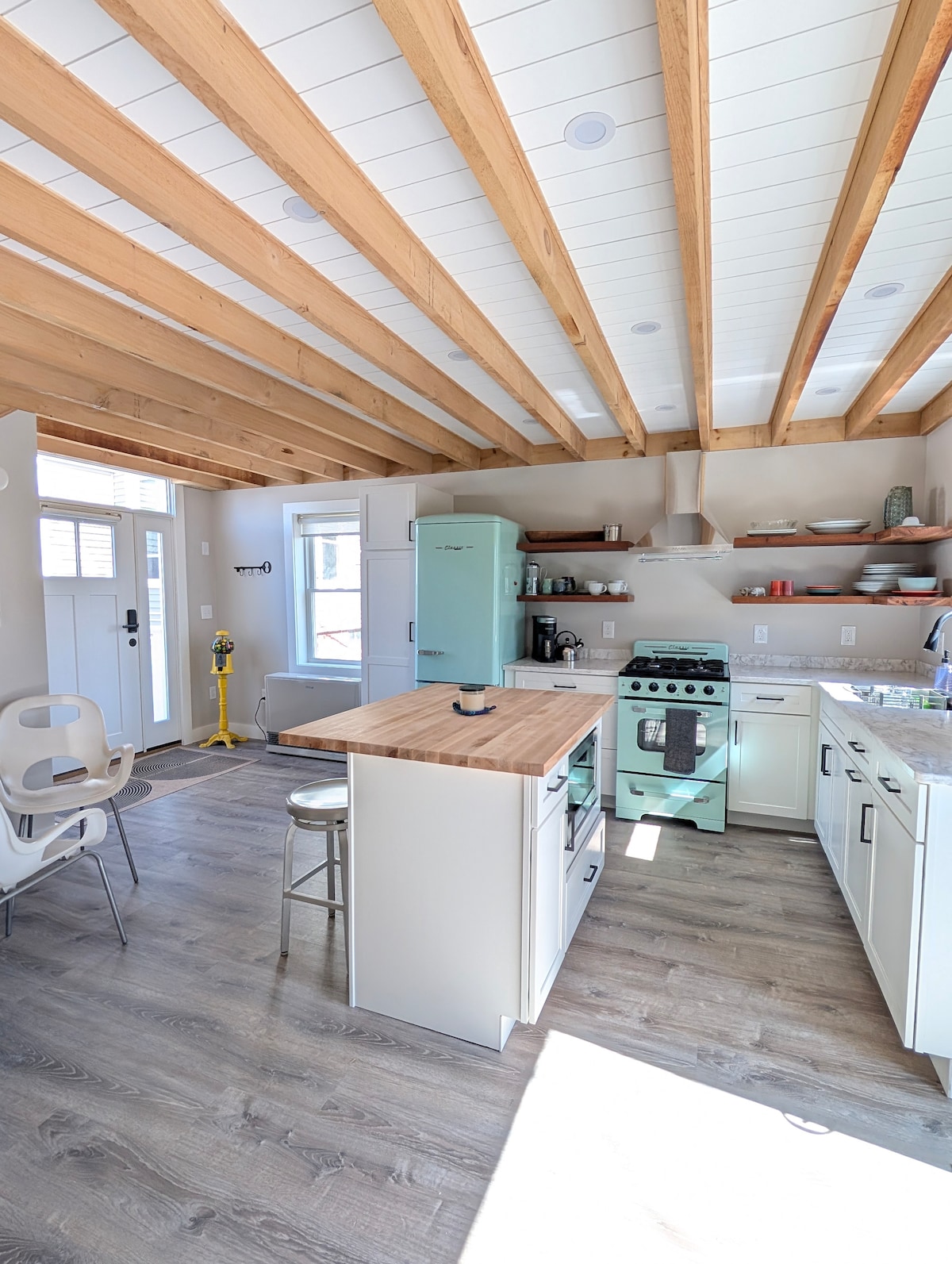 An open-concept kitchen features mint green retro appliances and a butcher block island with two bar stools. Light pours in through a window, highlighting the warm wood beams above. Shelves display a mix of dinnerware and kitchen essentials, creating an inviting space for cooking and dining.