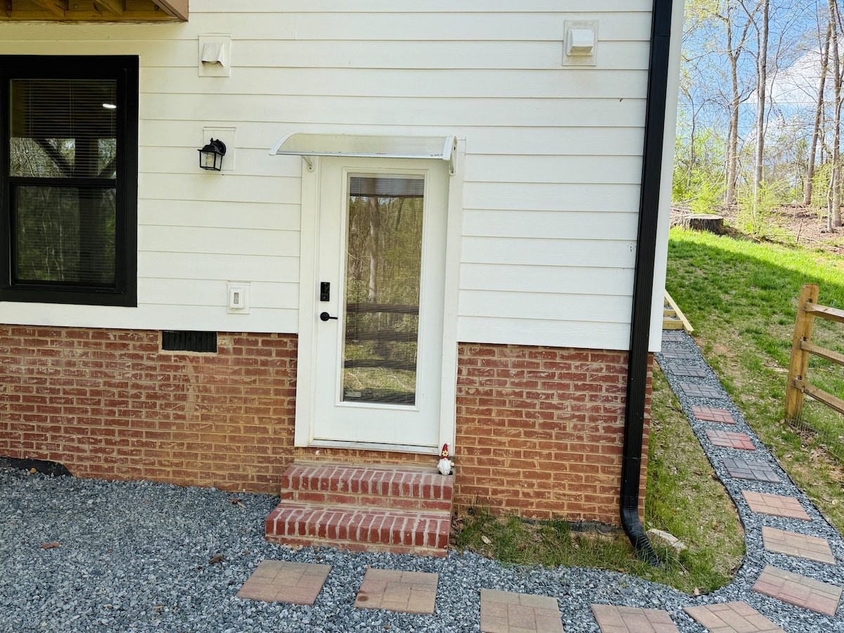 A private entrance is visible, featuring a glass door with a small step and an overhang for weather protection. Brick and stone pathways lead to the door, surrounded by lush greenery and a well-maintained yard.