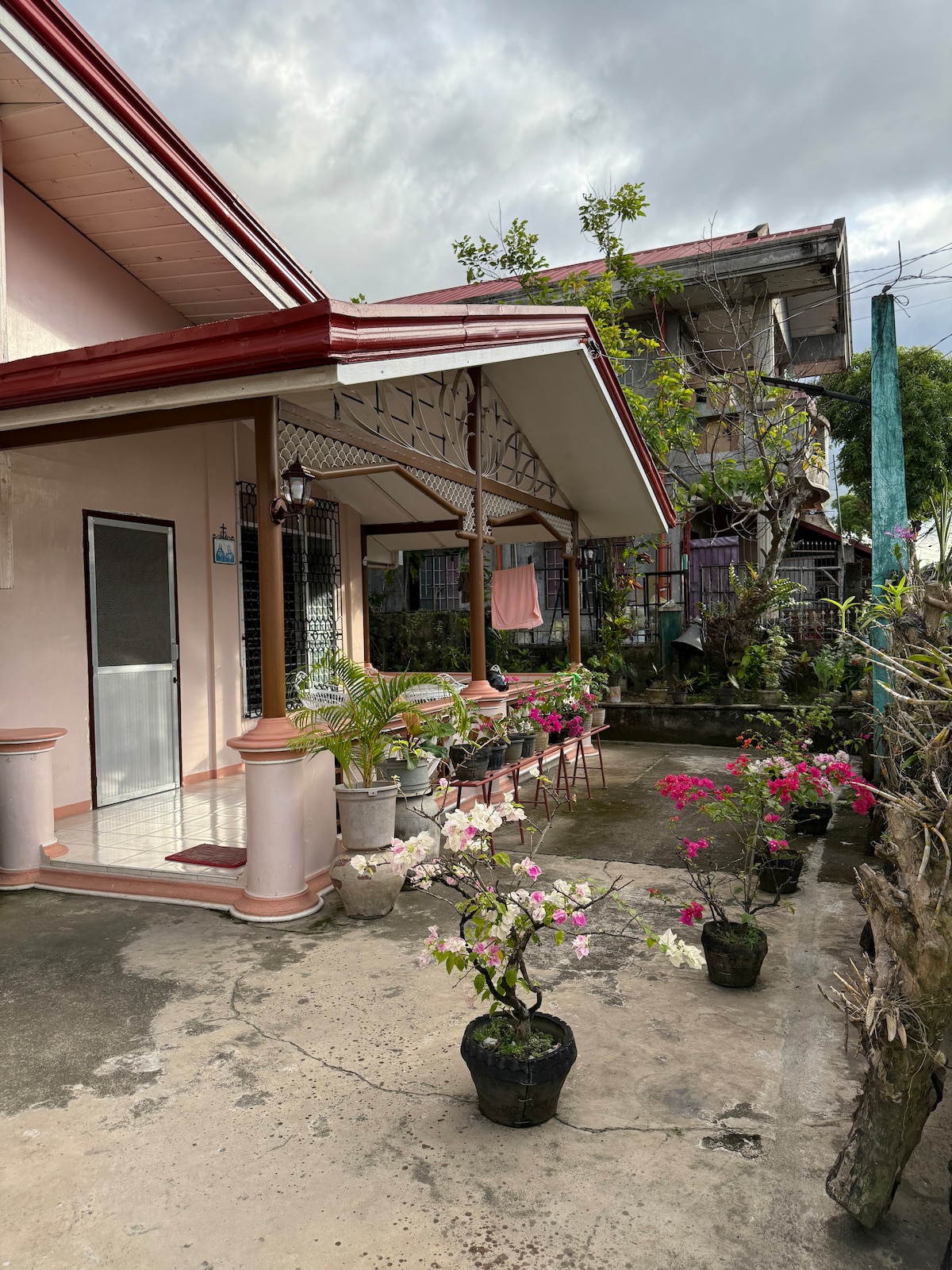 A charming exterior space is presented, featuring a covered terrace and potted plants. Several flowering pots line the pathway, adding color to the scene. The surrounding houses and greenery contribute to the inviting atmosphere of the garden area.