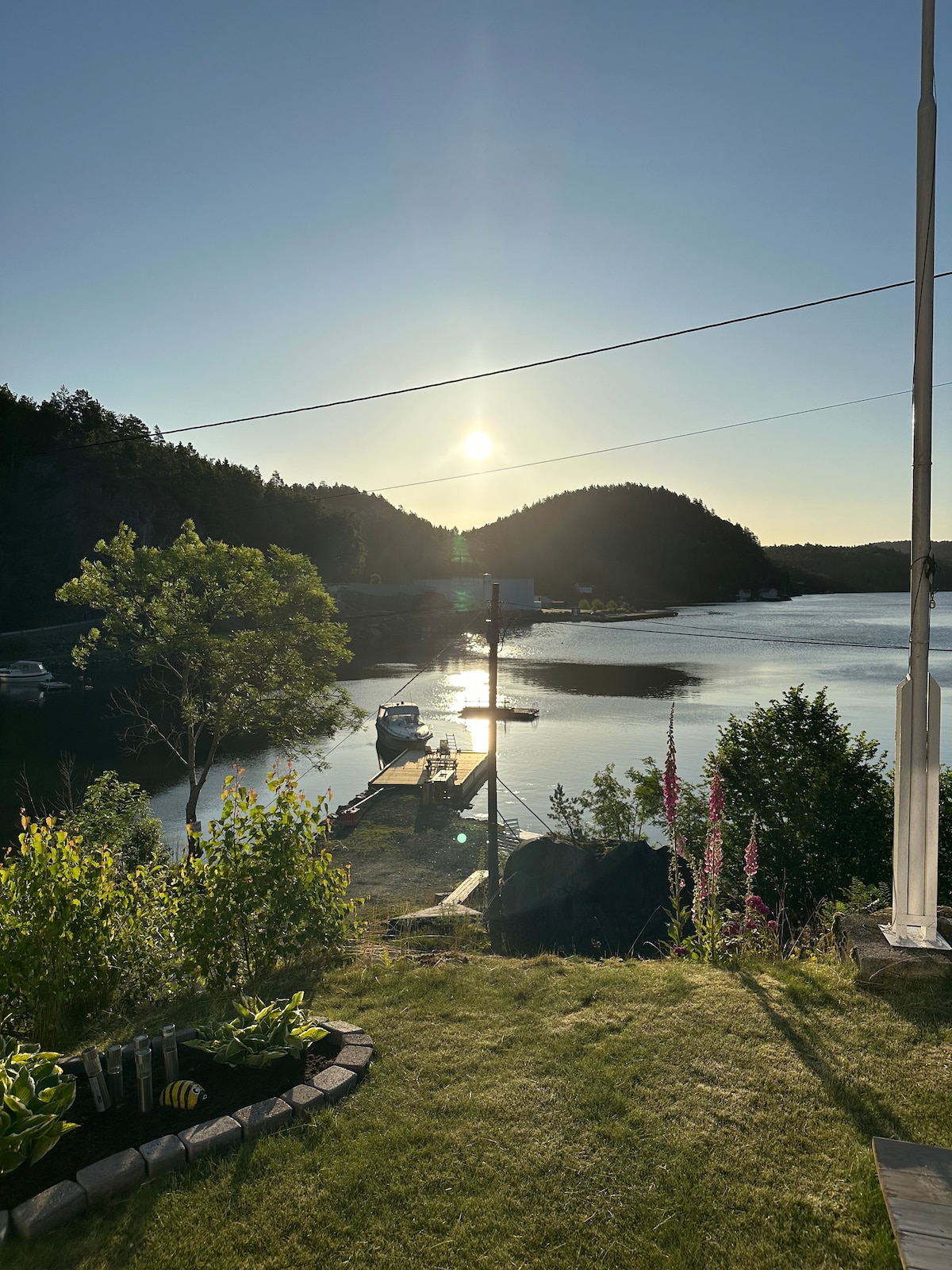 A scenic waterfront view captures the sun setting over the calm water of Fossingfjorden. Lush greenery lines the foreground, while a private dock extends into the water, with boats visible nearby. The surrounding hills add to the serene environment.