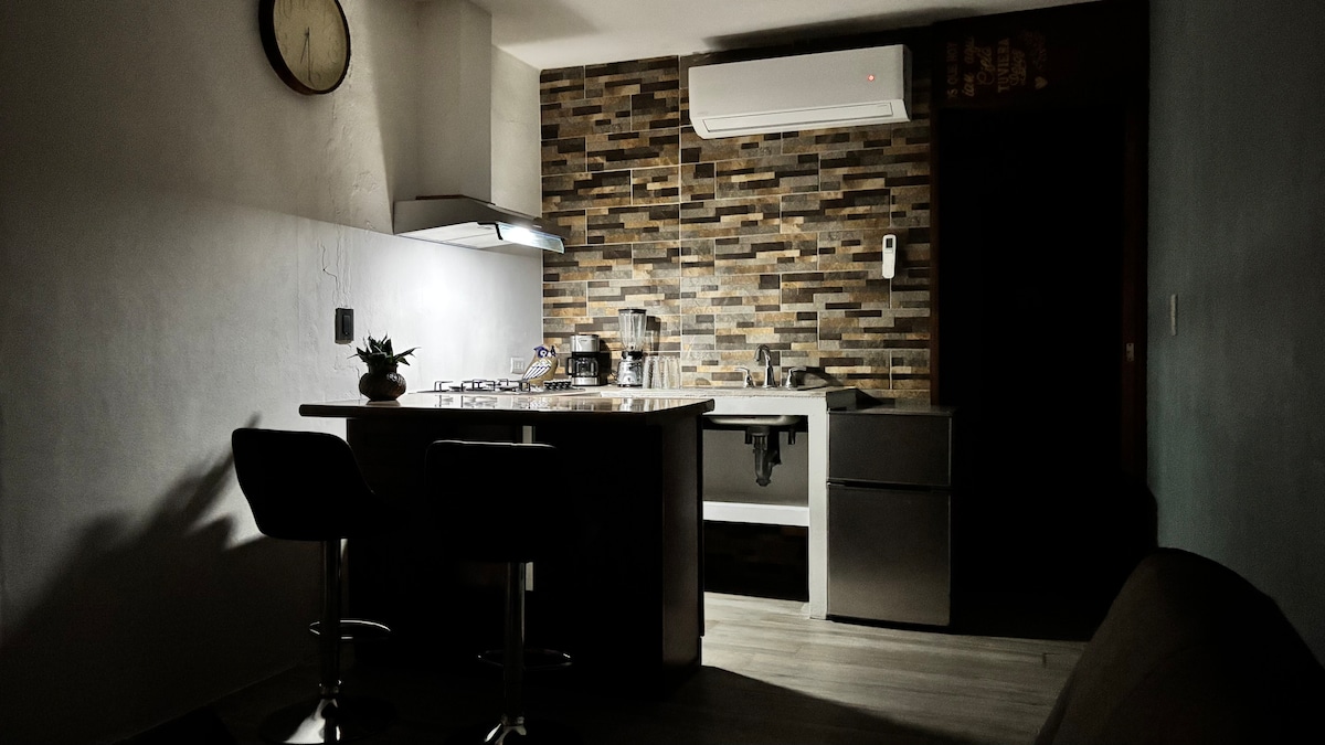 A kitchenette is visible, featuring a modern design with a stone-tiled backsplash. Two bar stools are positioned at a counter. A refrigerator, stove, and various kitchen utensils can be seen, along with a small potted plant on the countertop. Soft lighting creates a cozy ambiance.