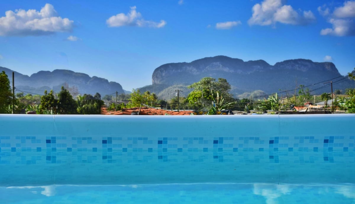 The image captures a serene view of a private pool with crystal-clear water, framed by vibrant blue skies and distant mountains. The lush greenery surrounding the area contributes to the tranquil atmosphere, inviting relaxation and connection with nature.
