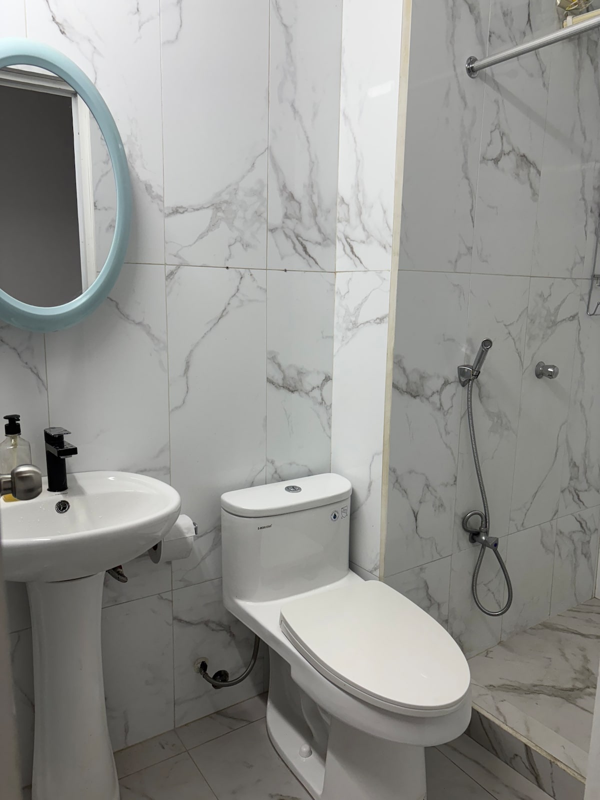 A modern bathroom is presented, featuring marble-patterned tiles in light tones. A round mirror with a pastel blue frame is mounted above a white sink. A toilet is placed adjacent to a walk-in shower equipped with a handheld showerhead and minimalist fixtures, offering a clean and spacious look.