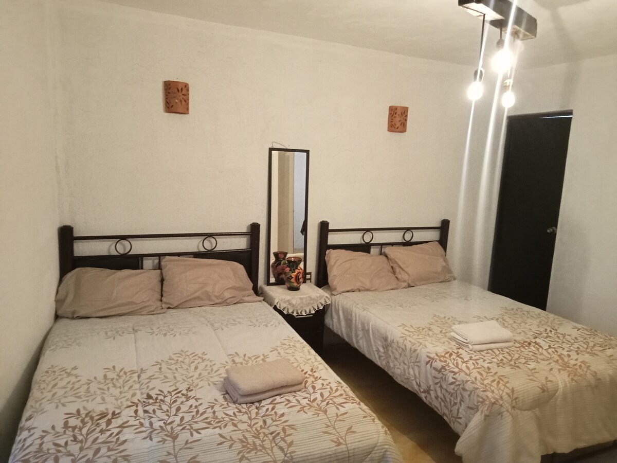 A dual sleeping area features two beds covered with light-colored linens. A small nightstand with a decorative vase is placed between the beds. A mirror reflects the soft light from pendant bulbs above, enhancing the overall brightness of the space.