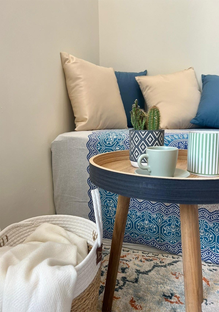 A seating area features a light gray cover with patterned accents, adorned with blue and neutral pillows. A low wooden table, holding a small potted plant and cups, stands beside a woven basket containing a throw. Natural light fills the space, enhancing its inviting feel.