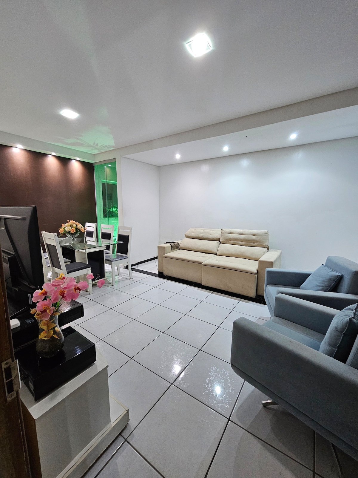 A bright living area features a beige sofa and a blue armchair, with a glass dining table set for four. The space is complemented by soft lighting and glossy tiles that enhance the room's contemporary design.