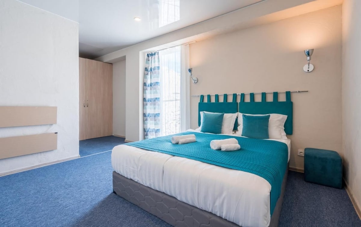 A spacious sleeping area features a large bed dressed in a bright blue coverlet, accented with cushion pillows. Natural light filters through a large window adorned with striped curtains. A nightstand with a lamp and a stool is also visible, along with a closet in the background.