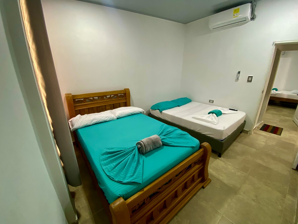 A well-lit sleeping area features two beds, one with a wooden frame adorned in a turquoise coverlet, and the other in a white coverlet. A towel is neatly arranged on the turquoise bed. Air conditioning is installed on the wall, contributing to a comfortable environment.