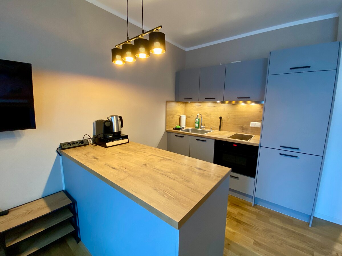 A modern kitchen area is showcased with sleek cabinetry in muted tones. A large countertop provides ample workspace, while a coffee maker and other kitchen essentials are neatly arranged. Soft lighting is installed above, enhancing the functionality of the space.