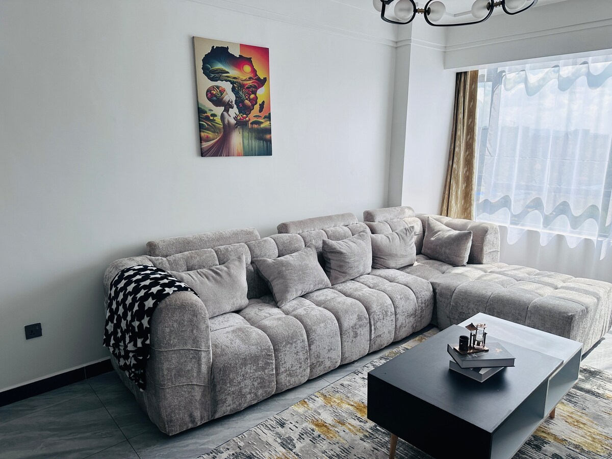A spacious living area features a large, comfortable gray sectional sofa adorned with a patterned throw blanket. A modern coffee table sits in front, complemented by a vibrant wall art piece. Natural light filters through a large window with sheer curtains, highlighting a neutral color palette.