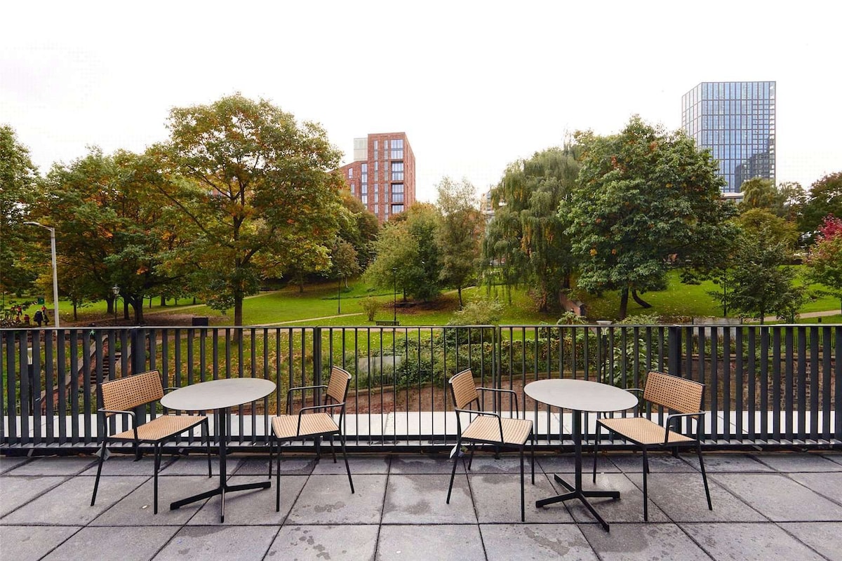 A spacious terrace is displayed with two round tables and four chairs, all positioned on a concrete surface. Lush greenery and trees are visible in the background, alongside urban buildings that provide a contrast to the natural scenery.