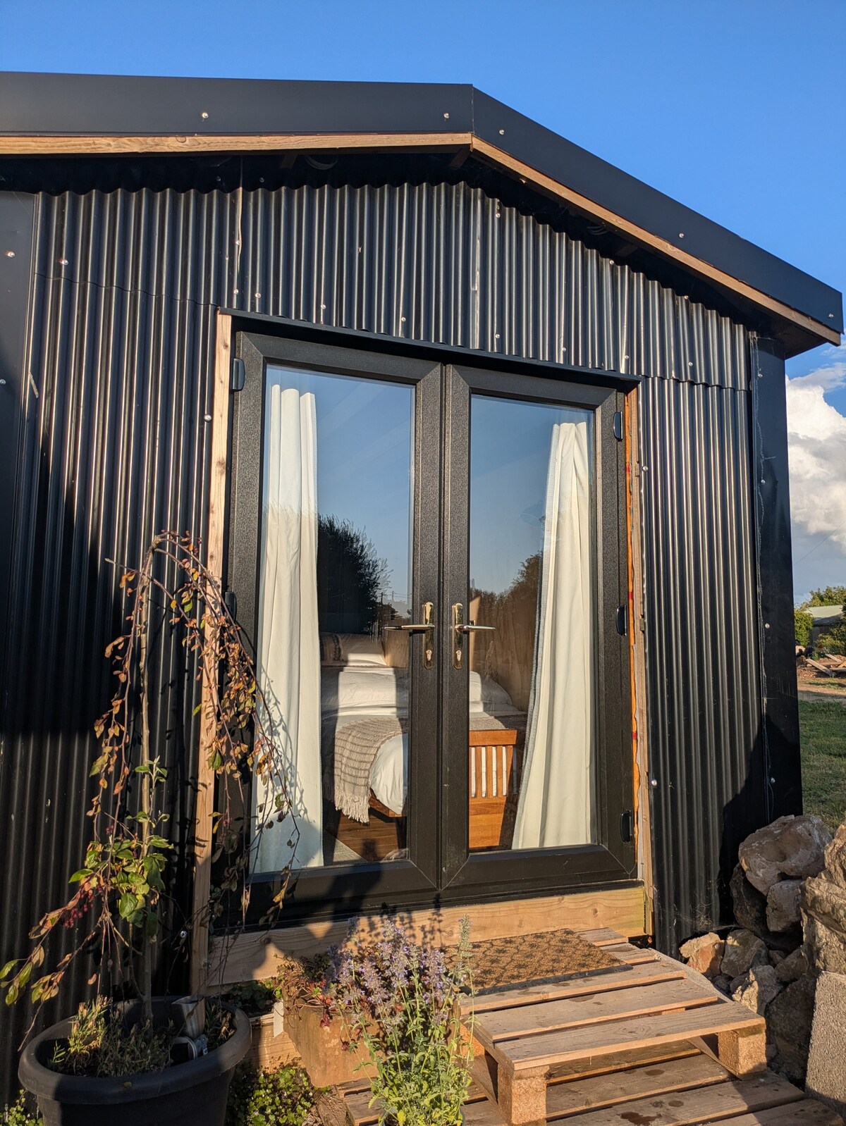 The exterior of the cabin is showcased, featuring a sleek black corrugated metal facade and large glass double doors. Light-colored curtains frame the doorway, which leads to the interior. Wooden steps lead up to the entrance, complemented by a small garden area with plants.