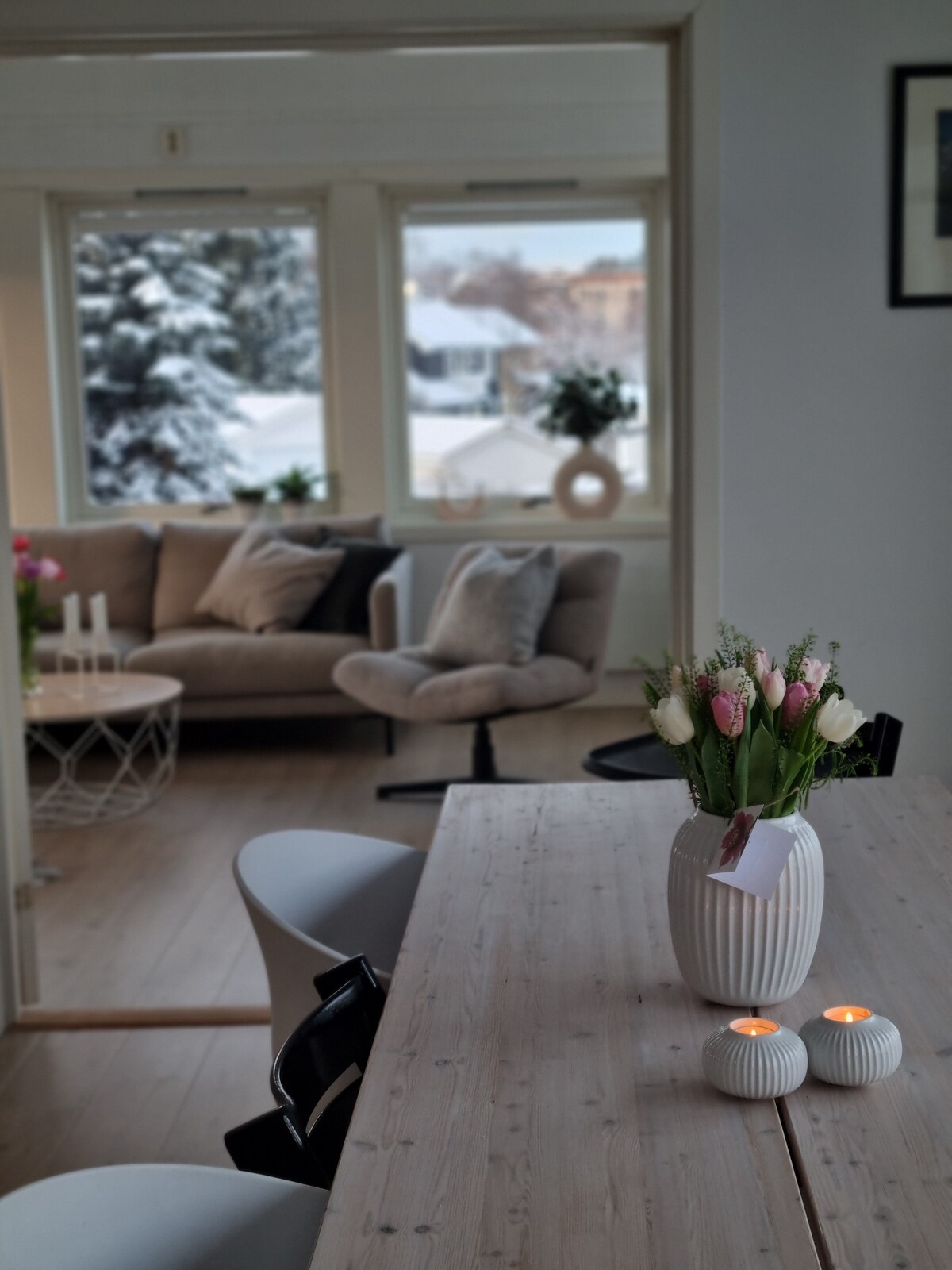 A bright dining area features a wooden table accompanied by black and white chairs. A vase filled with an assortment of flowers sits in the center, while two candles add a soft glow. A cozy seating arrangement with a sofa and armchair is visible through the doorway.