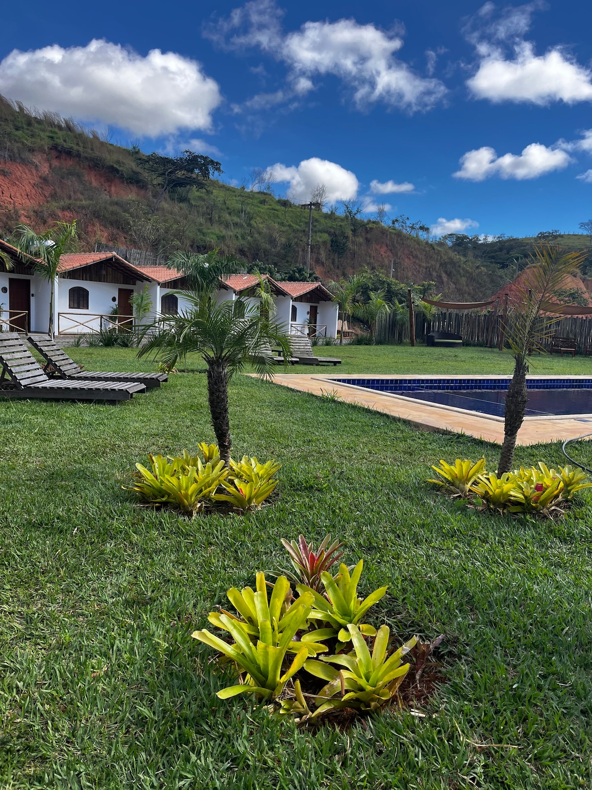 A serene outdoor area is presented, featuring well-maintained green grass and small tropical plants. A refreshing pool occupies one corner, with lounge chairs positioned nearby. Charming chalés are visible in the background, set against a backdrop of lush hills and a clear blue sky.