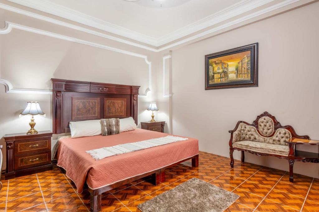 A spacious bedroom features a large wooden bed framed by nightstands with lamps. The polished floor complements the decorative artwork hung on the neutral wall, and a stylish seating area is included with an upholstered loveseat.