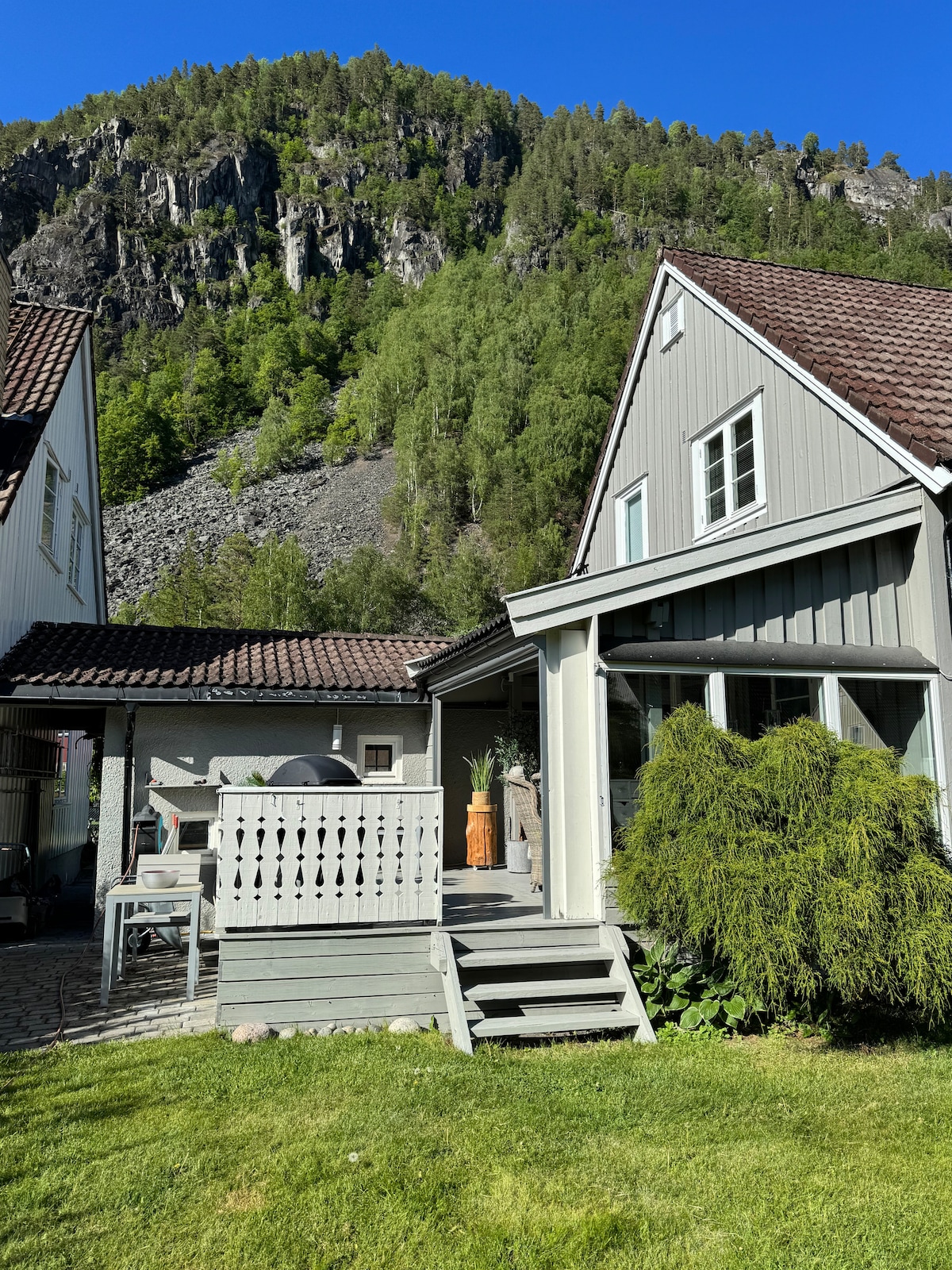 A charming exterior view displays a two-story house with a sloped roof. A wooden deck features outdoor seating and is surrounded by lush greenery. Nearby, a rocky hillside rises in the background under a clear blue sky.