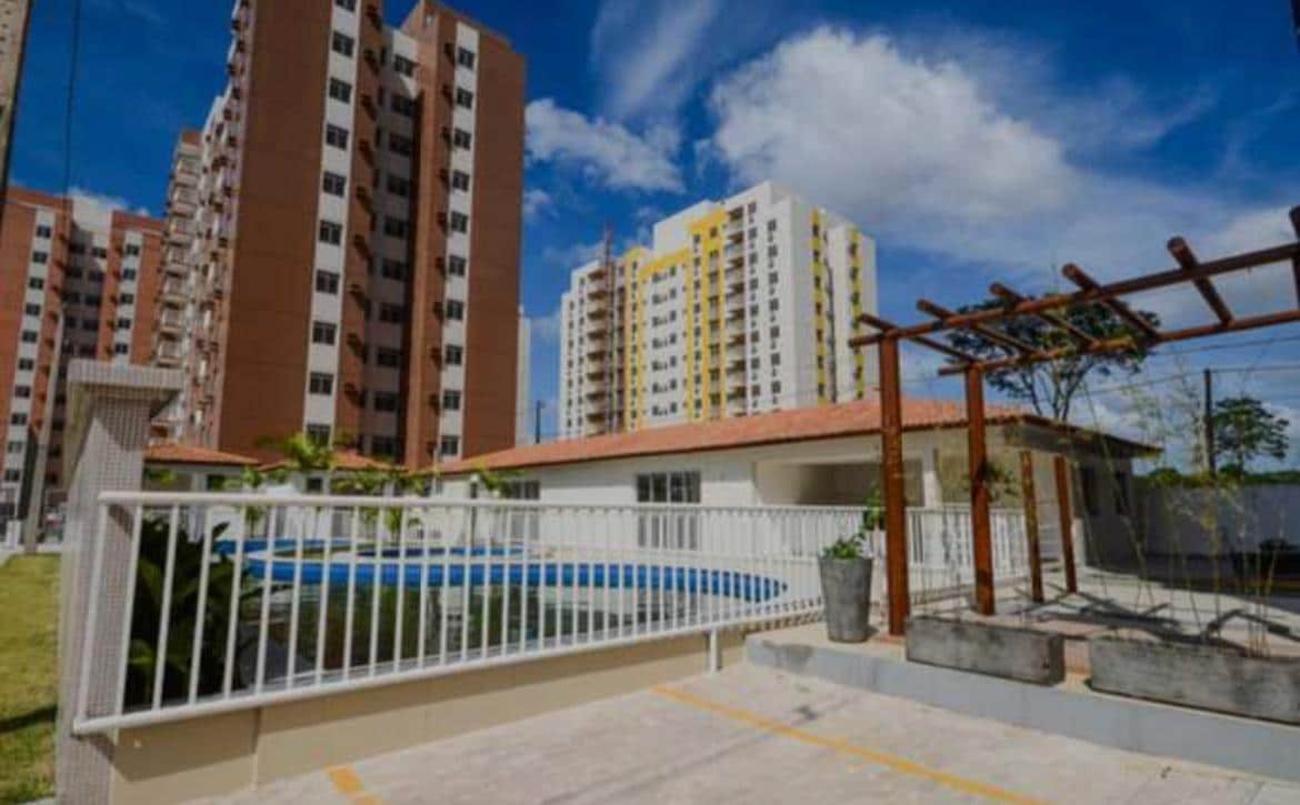 The image displays a landscaped area featuring an inviting swimming pool surrounded by a low fence. In the background, several multi-story residential buildings rise against a clear blue sky, with a pergola and planters in the foreground, enhancing the community atmosphere.