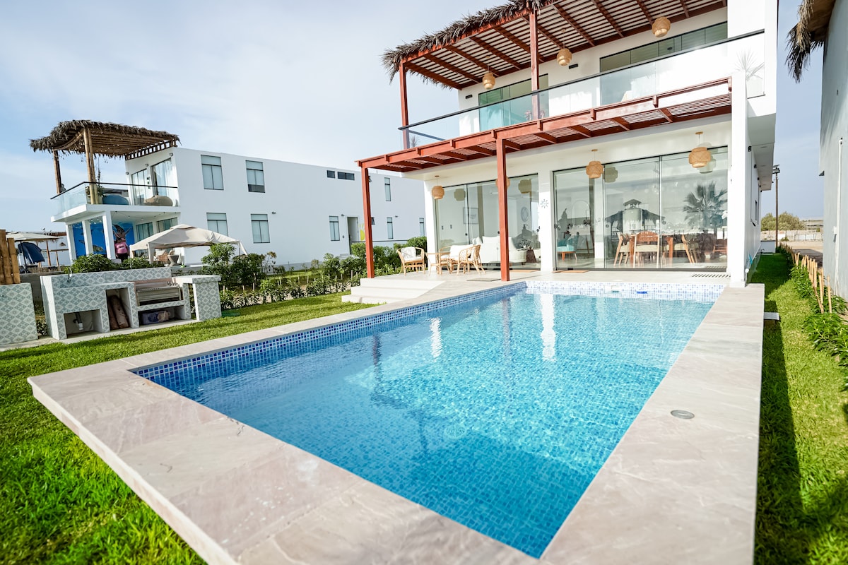 A private pool glistens under the sun, bordered by a spacious deck and manicured grass. The two-story home is visible in the background, featuring large glass doors that connect the indoor and outdoor living spaces.