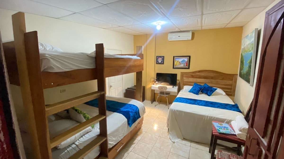 The room features a spacious layout with two beds; one is a wide bed and the other is a bunk bed with a lower twin and an upper twin. A small television and a desk are positioned between the beds, while artwork adorns the welcoming walls.