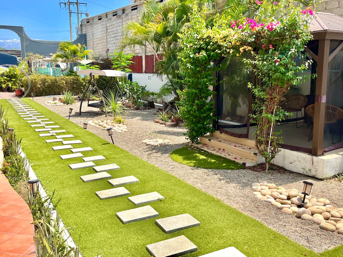 A landscaped garden area is shown, featuring a path of stepping stones leading through vibrant green grass. Tropical plants and flowers add color to the space. A covered gazebo is visible in the corner, providing a shaded seating area, enhancing the outdoor ambiance.