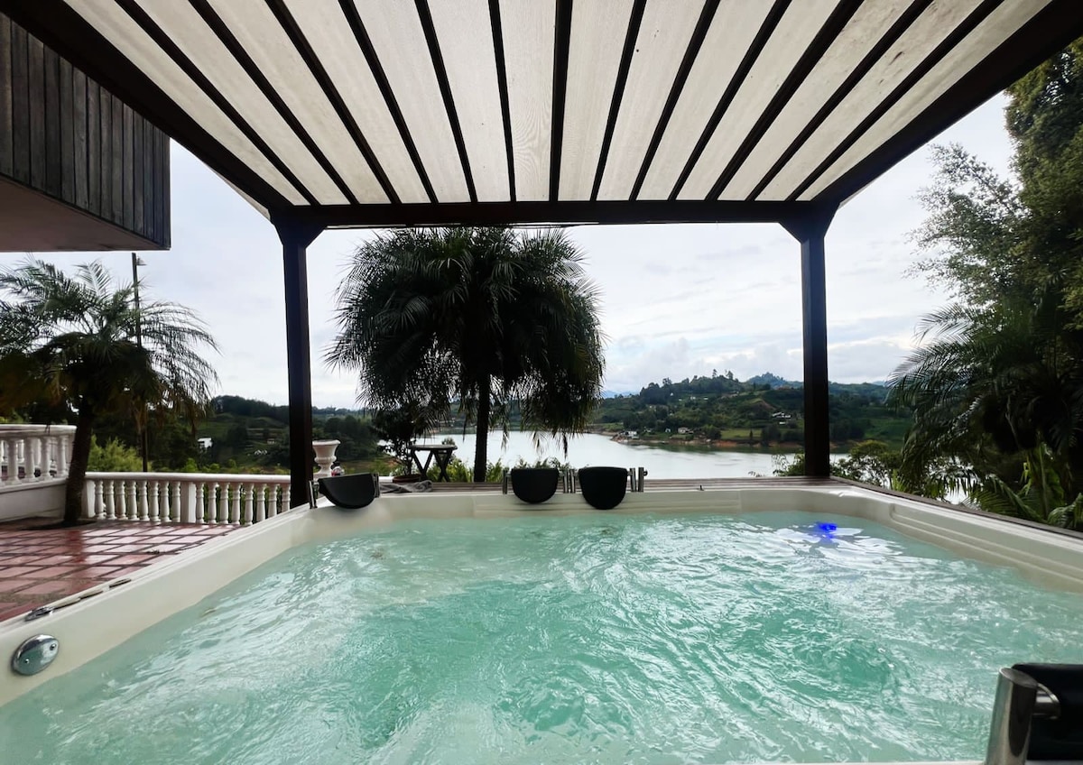 An outdoor jacuzzi rests under a slatted pergola, framed by lush greenery. The bubbling water reflects the sky and offers scenic views of the surrounding landscape, including rolling hills and a nearby body of water.