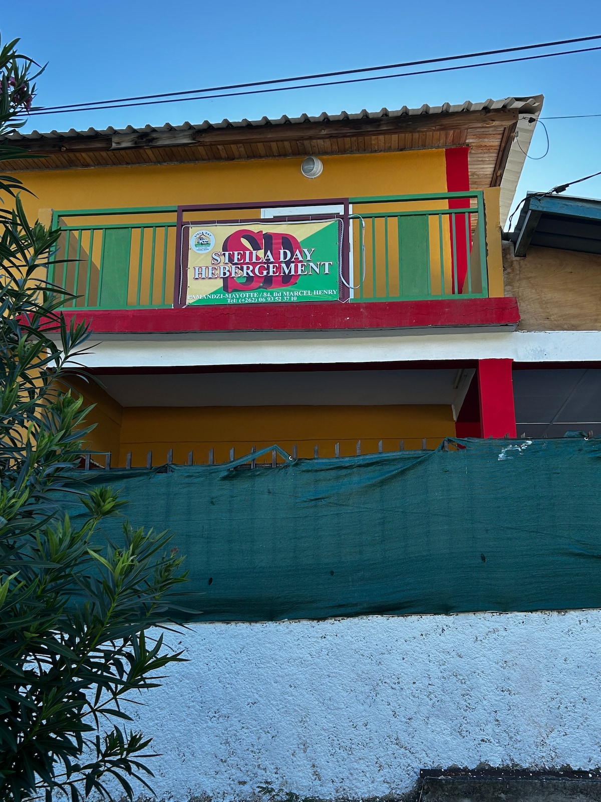 A colorful building exterior is shown, featuring a bright yellow wall with red and green accents. A sign indicating the name 'Stella Day Hébergement' is prominently displayed on the upper level, while greenery is visible in the foreground.