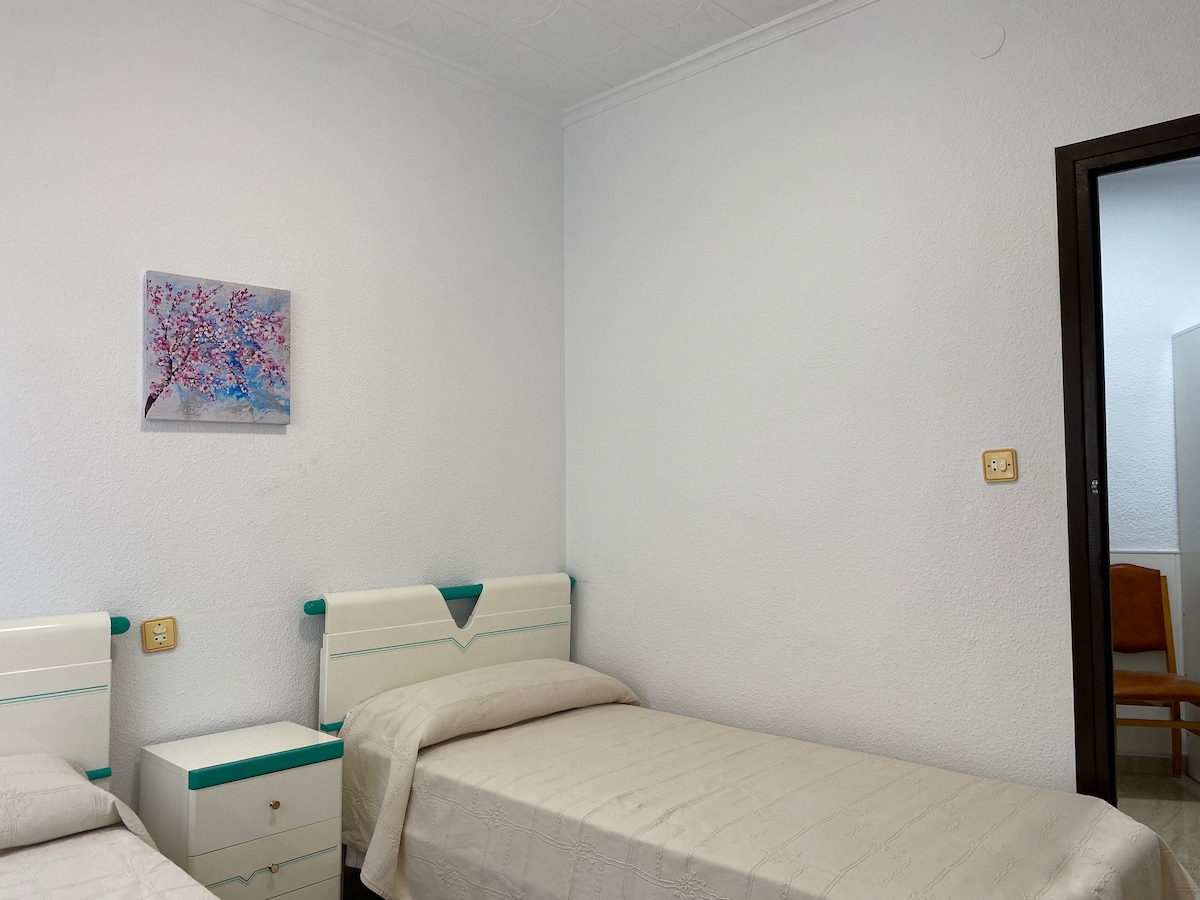 A well-lit room features two individual beds with light-colored bedspreads. Each bed is accompanied by a small bedside table. A piece of art depicting blossoms is displayed on the wall, and a doorway to another room is visible in the background.