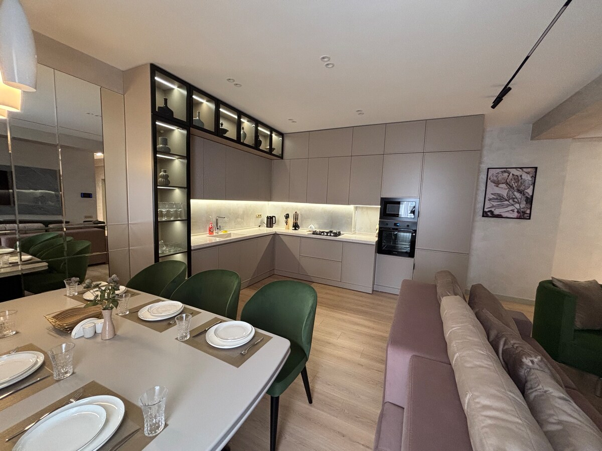 The kitchen area features modern cabinetry and high-quality appliances, including a built-in oven and stove. A dining table with five chairs is set for meals. Soft lighting highlights the sleek surfaces, and a comfortable seating area with plush sofas is visible in the background.