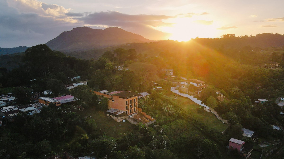 The image displays a hillside view during sunset, with the sun casting warm light over a landscape of lush greenery and scattered homes. A prominent building is positioned in the foreground, surrounded by trees and hills, creating a serene atmosphere of nature.