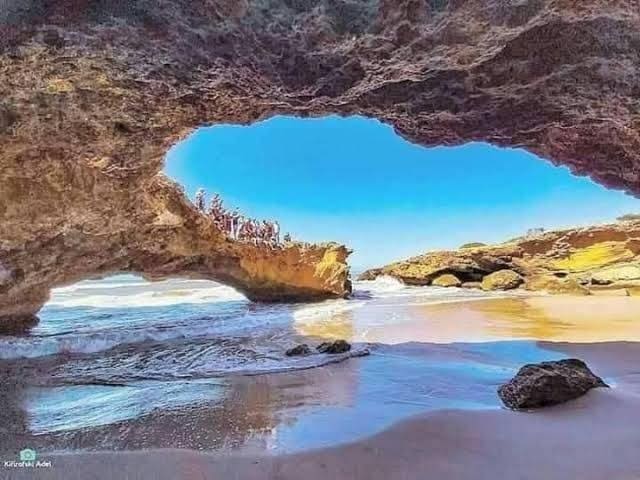 An expansive view of a rocky coastal area is framed by a natural arch. The sandy beach leads toward gentle ocean waves, while a clear blue sky is visible through the opening. Shadows and textures of the rocks are highlighted by the sunlight.